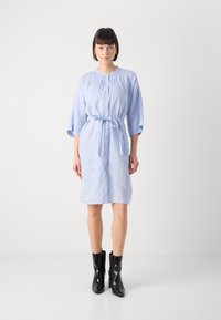 Svetlo modra lanena obleka s vezanim pasom, gumbi spredaj in širokimi rokavi, v kombinaciji s črnimi gležnjarji. Gladka tekstura, sproščen oblikovanje.