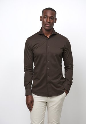Camisa de botones de manga larga color marrón, hecha de una tela suave. Presenta un cuello clásico y botones en la parte delantera. Combinada con pantalones de color claro.