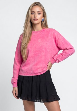 Unge kvinde med langt blond hår iført blå stjerneformede øreringe, en pink langærmet sweatshirt og et sort lagdelt skørt, stående med hånden på hoften.