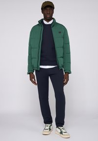 Zielona kurtka puchowa z wysokim kołnierzem, granatowy sweter, granatowe spodnie i zielono-kremowe sneakersy. Czapka w oliwkowej zieleni. Prosty, nowoczesny wygląd.