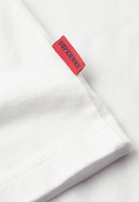 White cotton t-shirt with a red label featuring "SUPERDRY" stitched in blue, smooth texture, visible stitching along the seams.