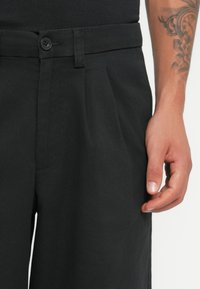 Black trousers made of smooth fabric, featuring a button closure, pleats, and side pockets. The waistband is flat and structured.