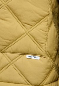 Gep quilteter senfgelber Jacke mit schräg verlaufenden Nähten, die ein sichtbares weißes Etikett mit der Aufschrift "MUSTANG" am Saum zeigt. Glatte Textur.