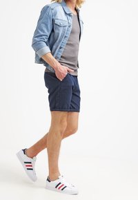 Farkkutakki, harmaa t-paita, tummansiniset shortsit, joissa on teksturoitu kuvio, sekä valkoiset sneakerit, joissa on punaiset ja siniset raidat. Vapaa-ajan asu liikkeessä.