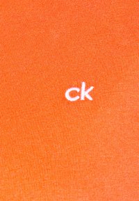 Oranžová bavlněná tkanina s jemnou ribkovou strukturou, s malým bílým vyšitým logem "ck". Jednoduchý design bez dalších vzorů nebo akcentů.