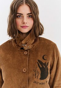 Brązowa kurtka z kordurowego materiału z wysokim kołnierzem, zapięciem na guziki oraz czarnym graficznym motywem wilka z drzewami i księżycem. Tekst brzmi "TU TYLKO DLA WYNAKLANIA."
