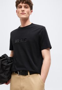 Νεαρός άνδρας με σγουρά μαλλιά, που φοράει μαύρο μπλουζάκι HUGO και μπεζ παντελόνι, κρατώντας μια μαύρη τσάντα με το ένα χέρι στην τσέπη.