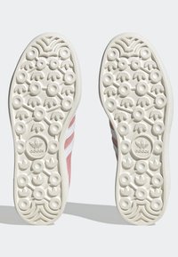 Par de suelas de goma blanca con patrones de dibujo circulares y triangulares, el logo de Adidas visible en el centro de cada suela debajo de unas zapatillas rosas.