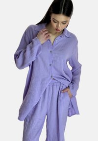 Camisa de botones lavanda y pantalones a juego en tejido texturizado. Corte holgado con cuello y botones frontales. Bolsillos visibles en los pantalones.
