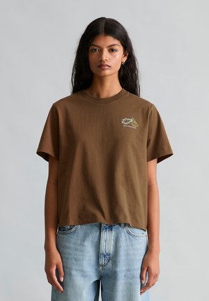 Frau mit langen dunklen Haaren trägt ein braunes T-Shirt mit Grafikdruck und hellblaue Jeans, steht vor einem einfarbigen grauen Hintergrund und schaut nach vorn.