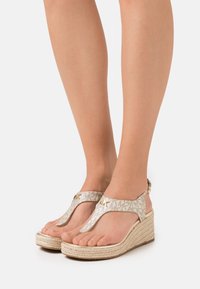 Sandali con zeppa caratterizzati da un upper in tessuto oro e beige con un motivo geometrico, un design a T e una suola dalla texture simile alla juta.
