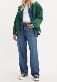 Zelená saténová bomber jacket s bílými pruhy, přes modrý denimový overal, spárovaná s širokými džínami a černými teniskami.