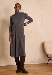 Maglione grigio con collo alto, abbinato a una gonna plissettata grigia e stivali marrone scuro al ginocchio. Gli accessori includono grandi orecchini ad anello.