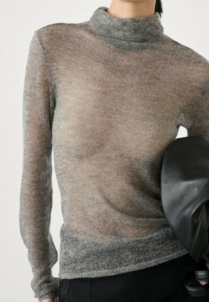 Person wearing a semi-sheer gray turtleneck sweater and holding a black leather bag under their arm.