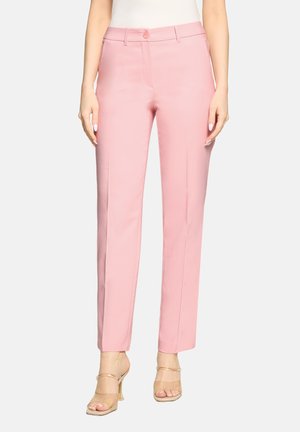 Frau trägt eng anliegende pinke gerade Hose mit Bügelfalte vorne, Gürtelschlaufen und nudefarbene Sandalen mit hohen Absätzen, steht vor weißem Hintergrund.