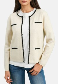 Cardigan en mélange de laine beige avec des bordures noires, col rond et deux poches avant. Assorti avec une chemise blanche et un jean bleu.