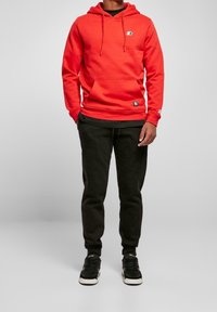Sudadera roja con bolsillo tipo canguro, cordón ajustable y logo en el pecho; combinada con pantalones de chándal negros y zapatillas negras.
