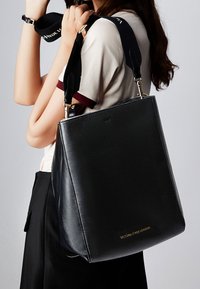 Černá kožená kabelka se strukturovaným designem, zlatým logem a odnímatelným černým popruhem. Hladký povrch, minimalistický styl.