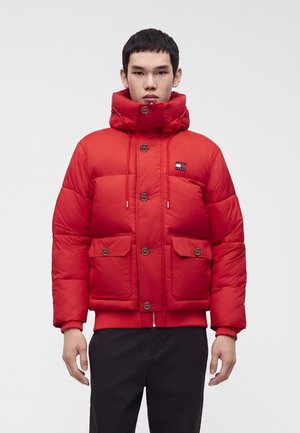 Un homme est tourné vers l’avant et porte une veste matelassée rouge vif, avec une capuche haute, des boutons sur le devant et de grandes poches.