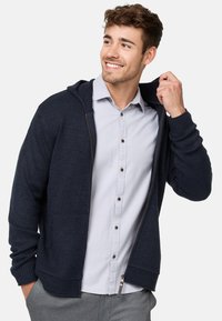 Navy knit hoodie with a zip front, ribbed cuffs, and a drawstring. Worn over a light gray button-up shirt with dark buttons.