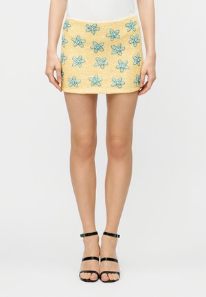 Yellow mini skirt with blue embroidered flowers worn with black strappy high-heeled sandals and a white top, shown from waist to feet.