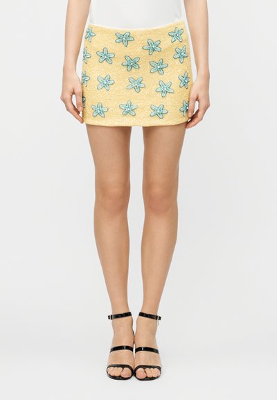 Gelber Minirock mit blauen, bestickten Blumen, kombiniert mit schwarzen, riemchenbesetzten High-Heel-Sandalen und einem weißen Oberteil, gezeigt von der Taille bis zu den Füßen.
