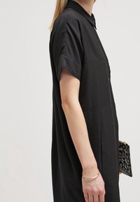 Abito camicia nero a maniche corte con colletto e tessuto testurizzato; tasche laterali visibili. Clutch nera a fantasia mantenuta in mano.