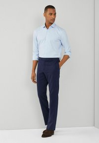 Camicia azzurro chiaro con bottoni abbinata a pantaloni blu marino dritti. Il modello sta in piedi con una mano in tasca, indossando scarpe in camoscio marroni.