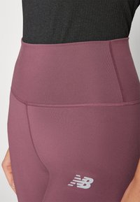 Legíny s vysokým pasem, pevně obepínající postavu, v tlumené purpurové barvě, s hladkým povrchem a bílým logem na dolní části nohy.