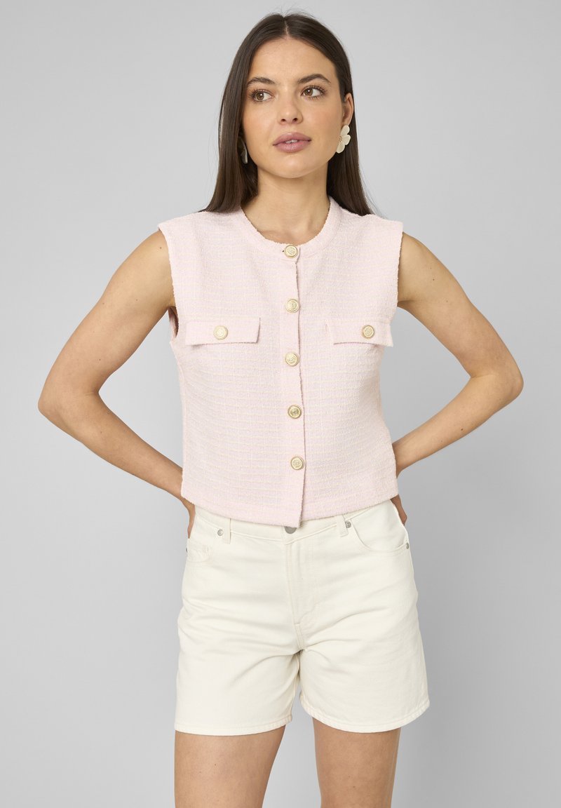 Femme aux longs cheveux foncés portant un gilet sans manches rose clair boutonné et un short écru, les mains sur les hanches, regardant légèrement vers la droite.