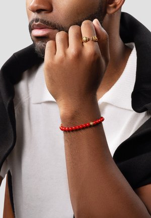 Rød perlearmbånd med guldfarvede accenter, der har glatte, runde perler og et enkelt design, båret på et håndled, mod en neutral baggrund.