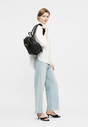 Vrouw gekleed in een wit overhemd, lichtblauwe spijkerbroek, zwarte sandalen met hakken, met een zwarte leren rugzak over één schouder gedragen.