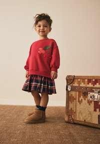 Roter Pullover mit Kirschdesign, karierte Röcke in Marineblau und Rot, braune Stiefeletten und kombiniert mit einem gemusterten Koffer. Neutrale Hintergrund.