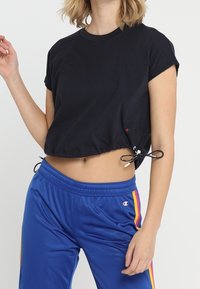 Černé zkrácené tričko s krátkými rukávy, s tkaničkou na lemu. Kombinováno s modrými sportovními kalhotami s vertikálními pruhy.