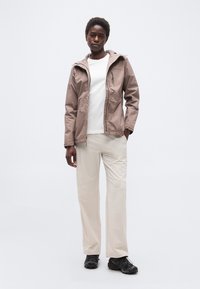 Άτομο που φοράει μπουφάν με κουκούλα σε χρώμα ταουπ, λευκό πουκάμισο, μπεζ παντελόνι και μαύρα παπούτσια πεζοπορίας, στέκεται μπροστά σε απλό φωτεινό φόντο.