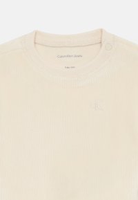 T-shirt pour bébé en coton côtelé de couleur crème avec un col rond, présentant un bouton-pression et un logo brodé "cK" à l'avant.