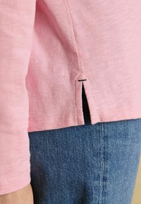 Lähedalpilt roosast pidadeseärkidega särgist külgsiludega ja musta õmblusdetailiga, kantud siniste teksade peal.