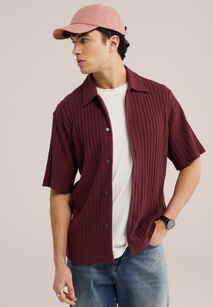 Junger Mann mit rosa Kappe, burgunderrotem, geripptem kurzärmeligem Hemd über weißem T-Shirt, blauen Jeans, schwarzer Uhr und Ring, der nach links schaut und die Hand in der Tasche hat.