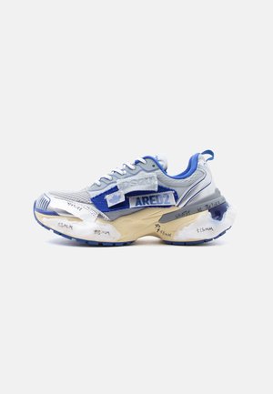 Zapatilla deportiva blanca y azul con parches de tela superpuestos, suela gruesa beige con amortiguación translúcida y notas de diseño escritas a mano.