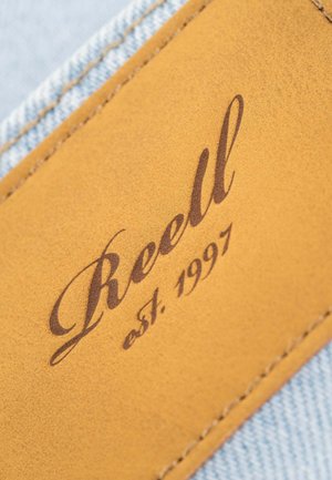 Parche de cuero con "Reell" en script marrón, con "est. 1997" debajo, adjunto a denim azul claro, mostrando textura y detalle de costura.