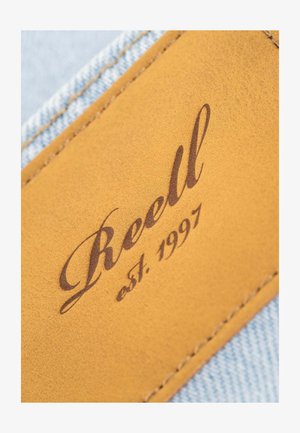 Parche de cuero con "Reell" en script marrón, con "est. 1997" debajo, adjunto a denim azul claro, mostrando textura y detalle de costura.