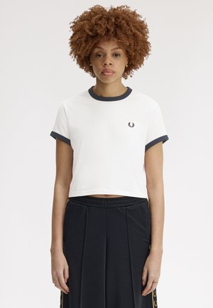 Femme aux cheveux bouclés portant un t-shirt court blanc avec une bordure noire et un pantalon noir plissé taille haute, debout devant un fond uni.