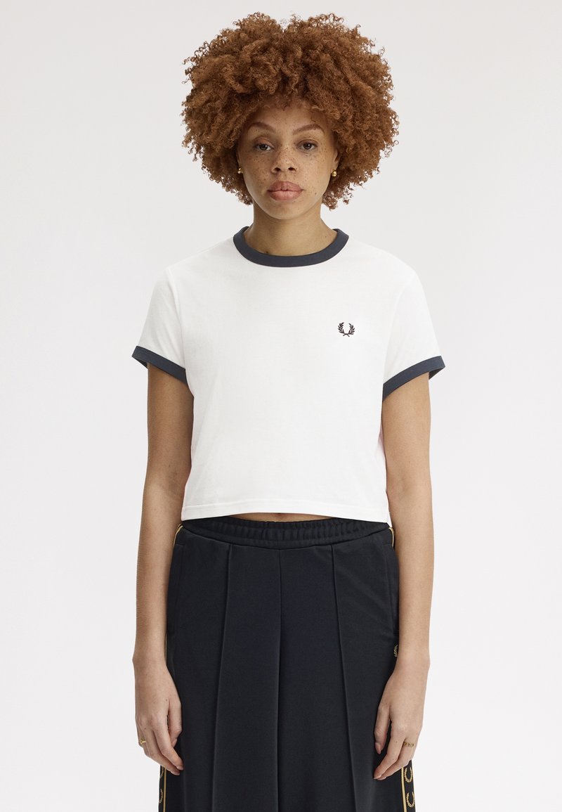 Femme aux cheveux bouclés portant un t-shirt court blanc avec une bordure noire et un pantalon noir plissé taille haute, debout devant un fond uni.