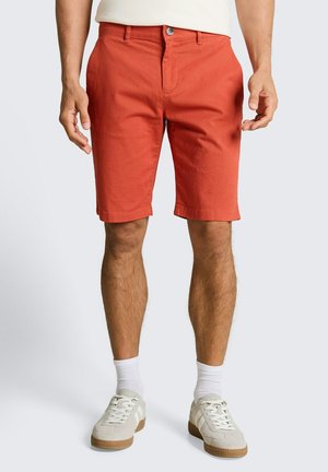 Man draagt roestkleurige knielange shorts, witte sokken en beige sneakers, staand tegen een effen lichte achtergrond.