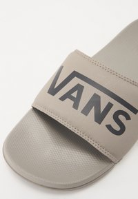 Sandale à glisser beige avec une large sangle comportant un logo "VANS" noir. Semelle intérieure texturée et semelle extérieure lisse avec une légère courbe.