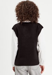 Veste noire en tricot avec manches courtes, ourlet côtelé et design au dos. Portée par-dessus une chemise blanche à manches longues, associée à un jean gris clair.
