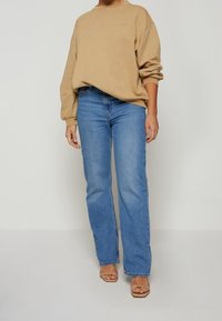 Sweat-shirt beige oversize avec manches retroussées, associé à un jean droit bleu clair. Porté avec des sandales à brides couleur tan.