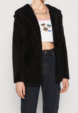 Personne portant une veste noire duveteuse, un crop top blanc avec des motifs papillons colorés, et un jean cargo taille haute bleu foncé.