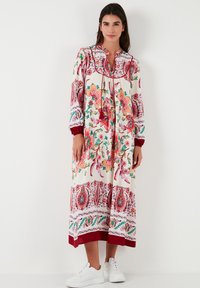 Vestido maxi floral con diseños vibrantes en rosa, verde y rojo. Presenta mangas largas, un collar y un dobladillo en capas. Combinado con zapatillas blancas.