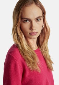 Fuchsia gestrickter Pullover mit Rundhalsausschnitt und weicher Textur, präsentiert an einem Model mit schulterlangem Haar und natürlichem Make-up.